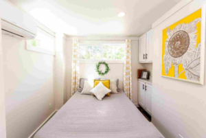 A bright, narrow bedroom with a single bed flanked by white cabinetry, yellow patterned curtains, a floral wreath on the wall, and abstract art.