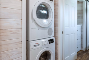 Stacked white washing machine and dryer in a modern laundry room with wooden walls and a white door.