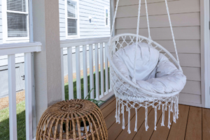 White hanging chair with cushions on a porch next to a small wicker table, with a backdrop of a house and fence.