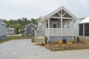 A small, modern modular home with a white porch and gray siding, numbered 58, located in a community with similar houses and green landscaping.