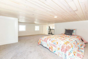 Spacious attic bedroom with slanted wooden ceilings, carpeted floors, and a bed covered in a colorful quilt.