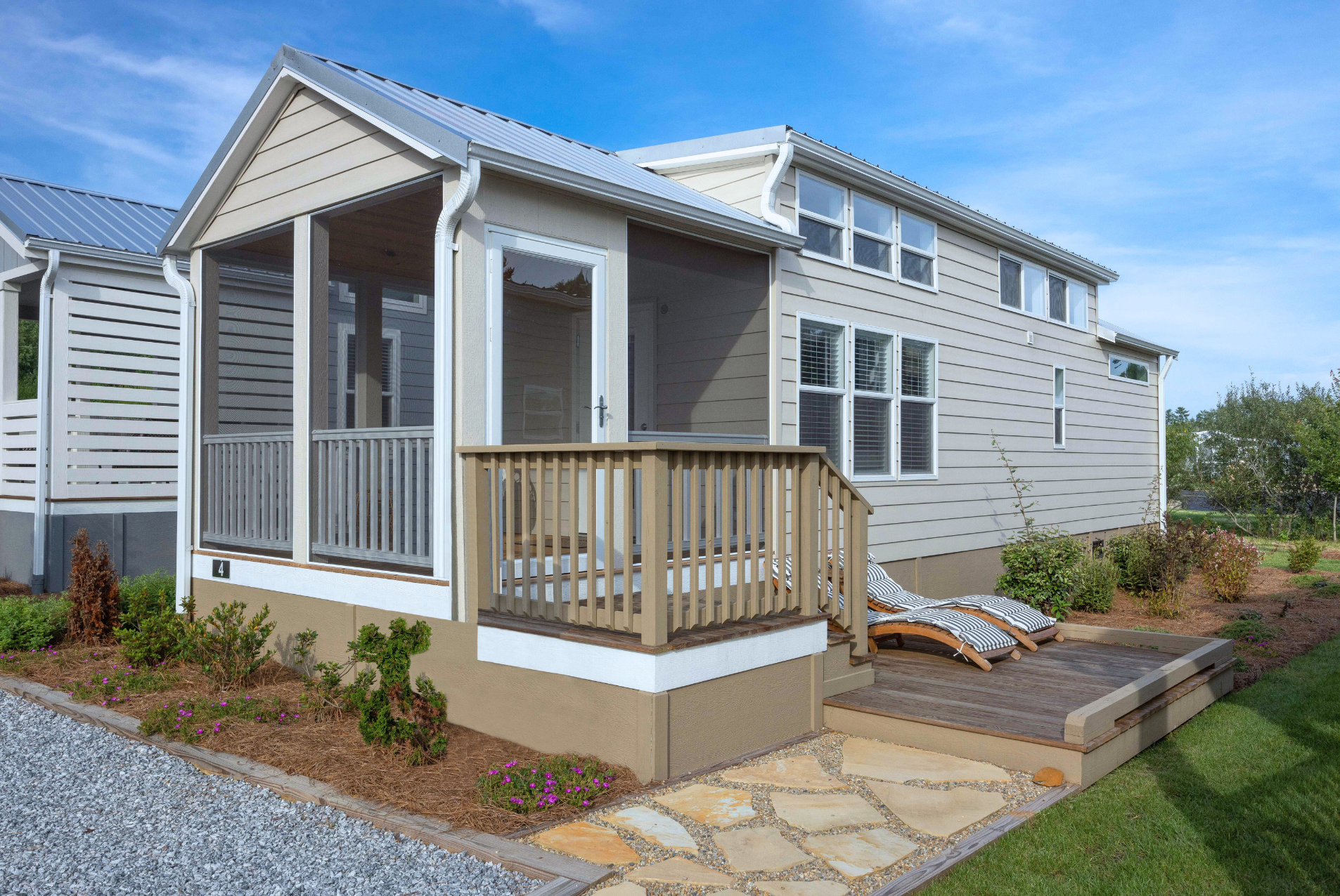 A modern tiny house with a wooden front deck, surrounded by a landscaped garden and clear blue sky.