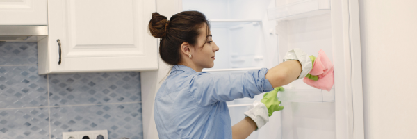 Woman in a blue shirt and green gloves cleaning the inside of an open refrigerator in a kitchen.