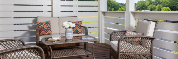 Outdoor patio with a wicker sofa, chairs, colorful pillows, and a small table with books and flowers, overlooking lush greenery.