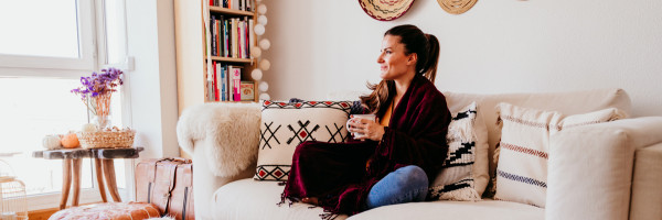 Woman sitting on a couch, holding a mug, in a cozy living room with bright decor and a small table with a pumpkin on it.