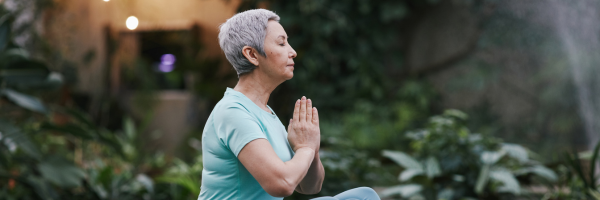 A senior woman with gray hair practicing yoga in a peaceful outdoor setting, performing a meditation pose with hands in prayer position.