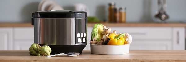 A modern multicooker next to a bowl of fresh vegetables on a kitchen counter.