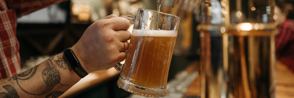 A person holding a frothy beer mug in a bar, with tattooed arms visible.