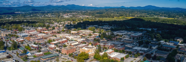Aerial view of a small town with dense buildings, surrounded by forests and mountains under a cloudy sky.