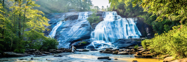 A cascading waterfall flows over rocks surrounded by lush greenery under a bright, sunlit sky.