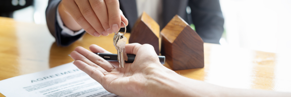 A real estate agent handing house keys to a client over a contract, with wooden house models on a desk.
