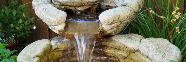 A stone garden fountain with water flowing from the top tier into a basin surrounded by lush green plants.