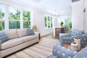 Bright, modern living room with a beige sofa, two patterned armchairs, and large windows overlooking green trees, connected to an open kitchen.