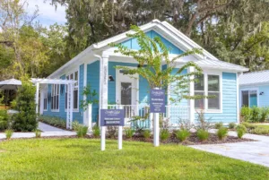 A charming light blue bungalow-style house with signs indicating it's a real estate agency, nestled under shady trees in a sunny setting.