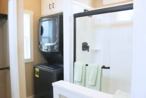 A modern laundry room with a black washing machine next to a glass-enclosed shower with green towels hanging on the door.