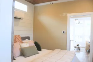 A cozy, minimalistic bedroom with a plush bed, soft pillows, wooden ceiling, and a door leading to a bright living area.