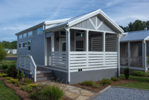 Modern tiny house with gray siding featuring a covered porch and wooden steps, set in a landscaped yard.