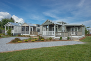 A modern, spacious modular home with gray siding and white trim features a large front porch and landscaped yard under a blue sky.