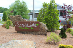 A landscaped entrance featuring a wooden sign reading "simple life the hamlet," flanked by large rocks and green shrubs under a cloudy sky.