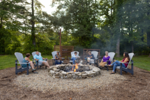 Group of people sitting around a campfire in a stone fire pit, chatting and relaxing in a wooded area during the day.