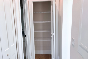 Empty walk-in closet with white open doors and multiple wire shelves, viewed from a hallway with wooden flooring.