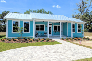 A single-story light blue house with a white front porch, metal roof, and a landscaped garden, under a clear sky.