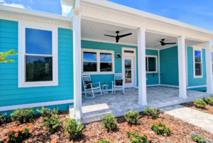 Bright turquoise house with a large covered porch, white rocking chairs, ceiling fans, and surrounded by landscaped garden.