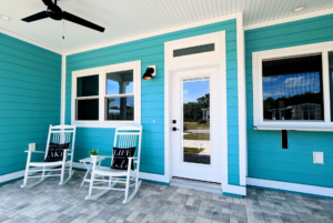 Bright blue house exterior with a white door, two white rocking chairs, and three windows. a ceiling fan hangs above on a covered porch.