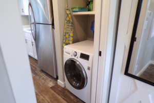 A modern laundry area with a front-loading washing machine beside a stainless steel refrigerator, set in a narrow space with white cabinetry.