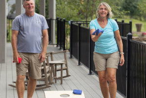 A senior man and woman playing a game of cornhole on a deck, smiling and holding bean bags.