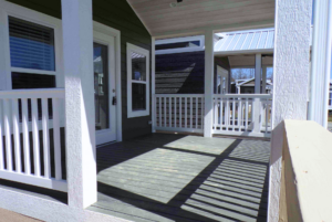 A sunny porch with white railings, a few steps, and shadow patterns on the wooden floor, leading to a house with a white door and windows.