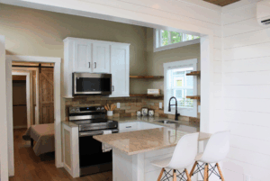 Modern kitchen with white cabinets, a small island, two chairs, and a view into a bedroom with a sliding barn door.