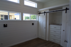 A modern room featuring white walls with a sliding barn door, built-in drawers, and wooden flooring. large windows provide natural light.