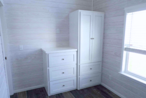 White wooden furniture consisting of a tall cabinet and an attached chest of drawers in a room with light wood paneling and a window.