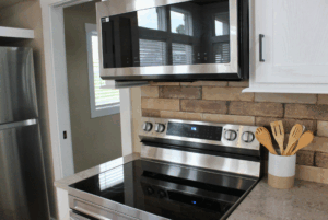 Modern kitchen with stainless steel oven, microwave, and a utensil holder on a countertop near a brick backsplash.