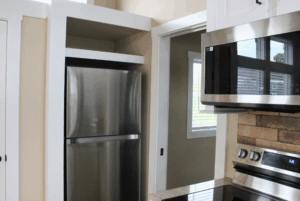 Stainless steel refrigerator in a kitchen nook, next to a microwave and oven, with a window in the background.