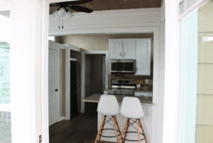 Modern kitchen with white cabinets, two white barstools, and stainless steel appliances, viewed through a doorway.