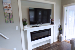 A living room corner featuring a wall-mounted tv above an electric fireplace, with decorative items on a small shelf and a wall art on the left.