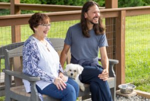 A man and a woman smiling and sitting on a bench outdoors with a small white dog on the man's lap. a water bowl is on the ground beside them.
