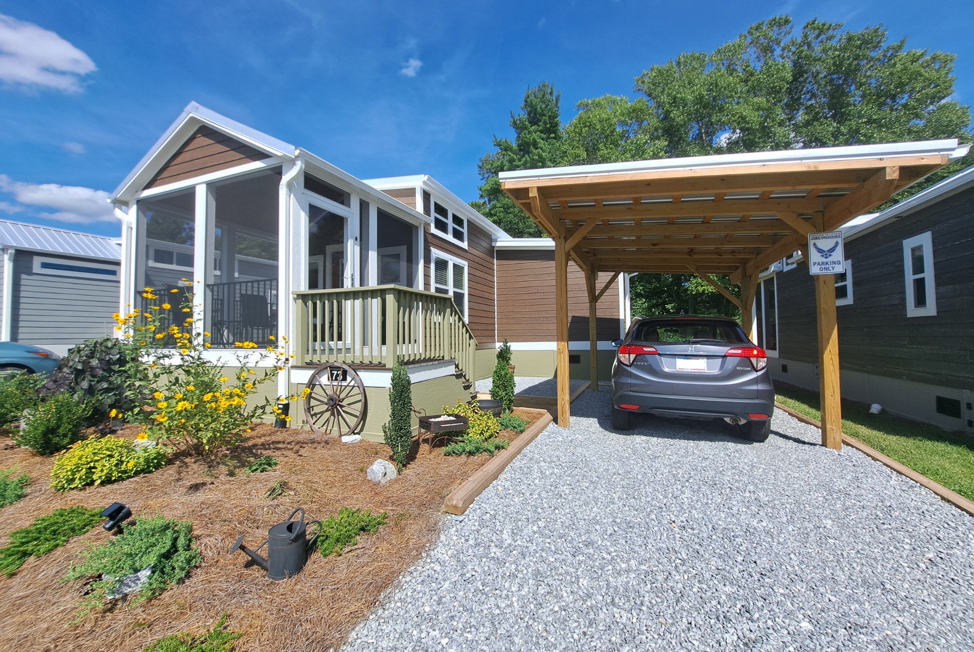 A small house with a white porch, a gravel driveway, and a carport. A car is parked beneath the wooden carport. The garden in front has yellow flowers and evergreen shrubs.