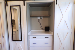 A modern small walk-in closet featuring a built-in white cabinet with drawers, a shelf with cables, and a mirror on the left wall.
