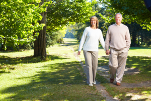 An elderly couple smiling while walking hand in hand on a sunny park path surrounded by trees and green grass.