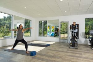 Two people exercising in a bright room with large windows. One person is doing yoga on a mat, and the other is on an elliptical machine. Various gym equipment and mirrors are in the background.