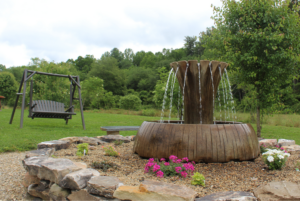 A rustic wooden barrel fountain with water spilling over in a landscaped garden, surrounded by rocks and flowers, with a swing bench nearby.