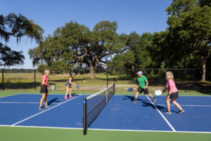 Four adults playing doubles pickleball on an outdoor court, surrounded by trees under a clear blue sky.
