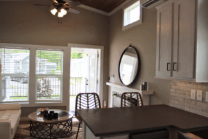 Modern living room with open door, round mirror, bar stools, and sunlight streaming from large windows into 2 beds.