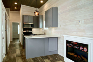 A narrow, modern kitchen with gray cabinets, white countertops, stainless steel appliances, and wooden walls. An electric fireplace is built into a white frame on the right side.