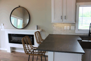 Modern kitchen with dark countertop, two woven chairs, round mirror, white fireplace mantel, and a subtle Swayback style.