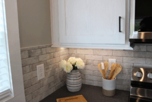 White roses swayback in a striped vase and wooden utensils on a kitchen counter with gray tile backsplash.