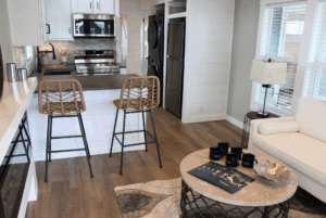Modern kitchen and living area with 2 beds, bar stools, white sofa, round table, and stacked washer-dryer.